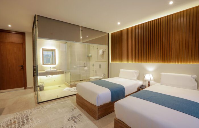 Modern hotel room with two twin beds, wood-paneled wall, and a glass-enclosed bathroom