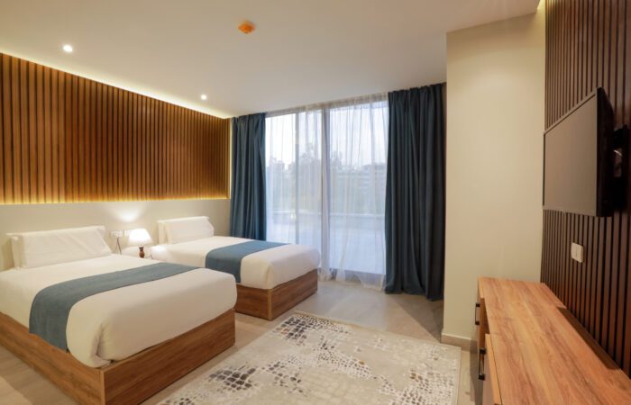 Modern hotel room featuring two twin beds, wood-slatted wall panels, and a large window with blue curtains