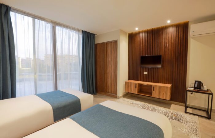 Modern hotel room with two twin beds, large window, and wall-mounted TV on a wood-paneled accent wall