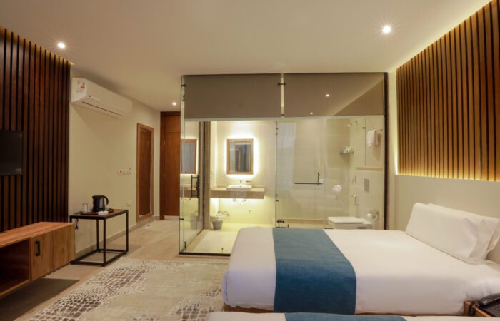 Modern hotel room with twin beds, wood-paneled walls, and a glass-enclosed bathroom
