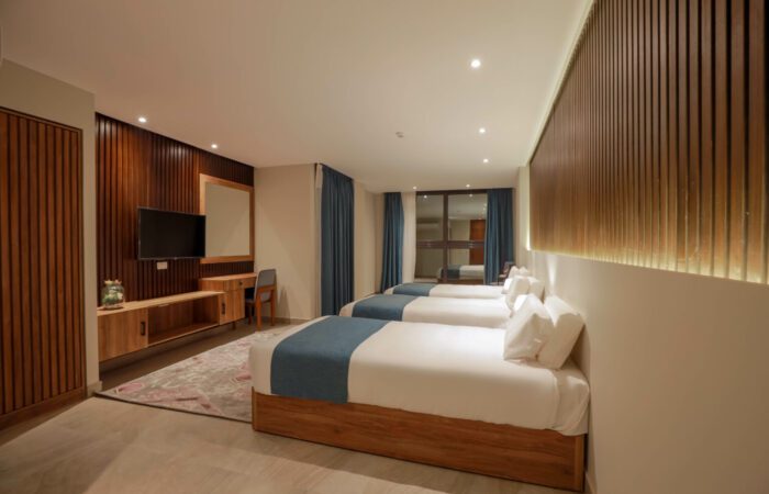 Modern hotel room featuring three single beds with blue runners, wooden wall paneling, and a wall-mounted TV
