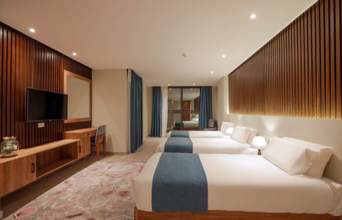Modern hotel room with three single beds, wooden accent walls, a desk, and a wall-mounted TV