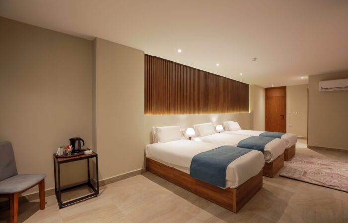 Modern hotel room featuring three single beds with white linens, blue runners, and a wooden slat feature wall