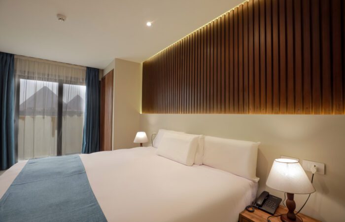 Modern hotel room with a white bed, wood-paneled wall, and a view of the Giza Pyramids through the window