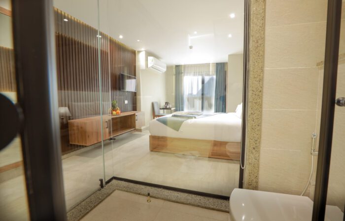 Modern hotel room with a large bed and TV, viewed through a glass partition from the bathroom