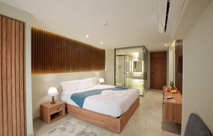 Modern hotel room with a large bed, wooden slat headboard, and a glass-enclosed bathroom