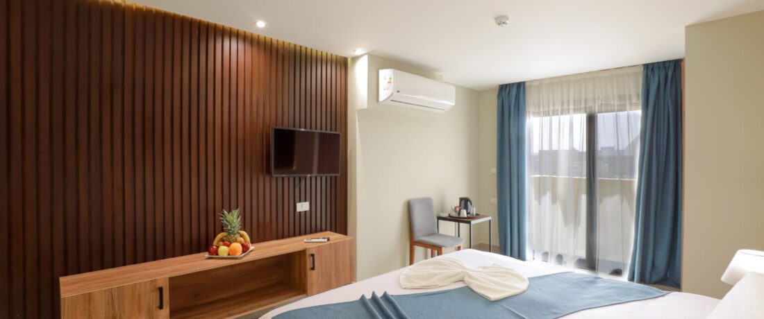 Modern hotel room with a bed, wood-slatted accent wall with a TV, and a window with blue curtains