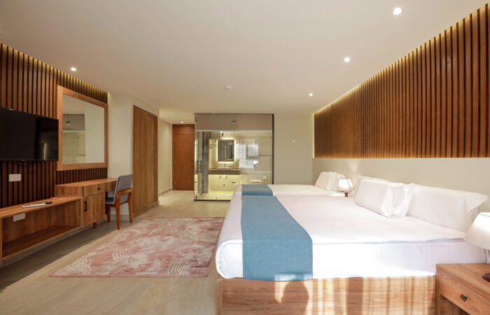 Modern hotel room with two beds, wood-slat accent walls, a desk area, and a glass-enclosed bathroom