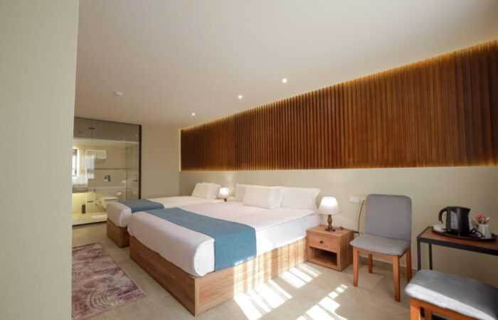 Modern hotel room with a double and single bed, wooden slat feature wall, and a glass-walled bathroom