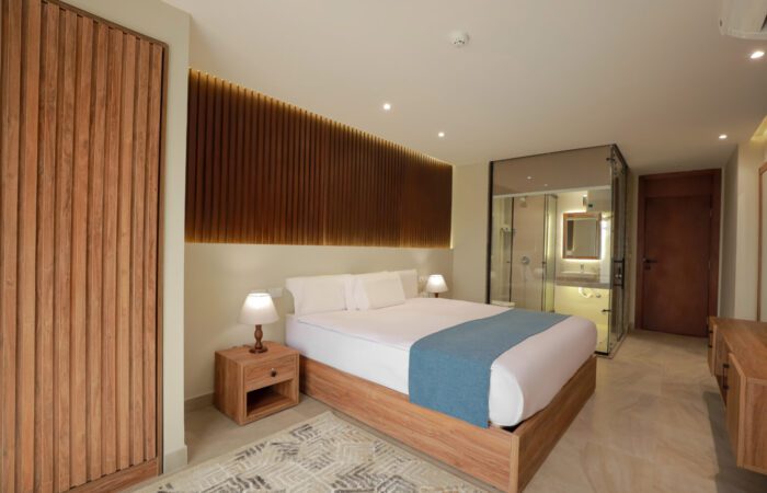 Modern hotel room with a wooden bed frame, slatted wood wall feature, and glass-enclosed bathroom