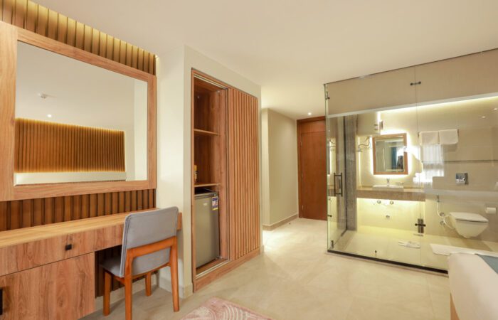 Modern hotel room featuring a wooden desk, a mini-fridge in a slatted closet, and a glass-walled bathroom
