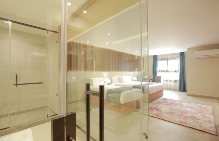 Modern hotel room with two twin beds and a glass-walled bathroom featuring a walk-in shower