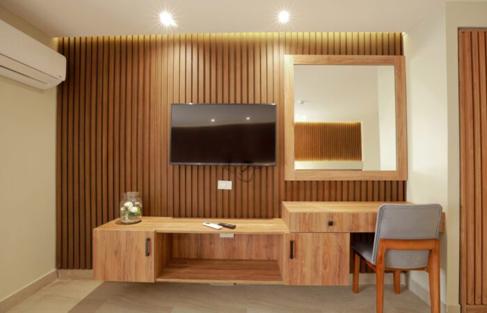 Modern room interior with a wood-paneled wall, wall-mounted TV, floating desk, large mirror, and a grey chair