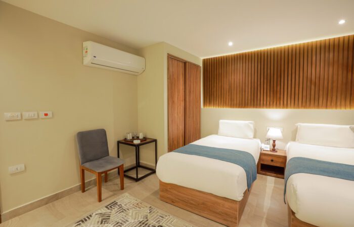 Twin hotel room with two single beds, wooden slat headboard, air conditioning, and a side chair