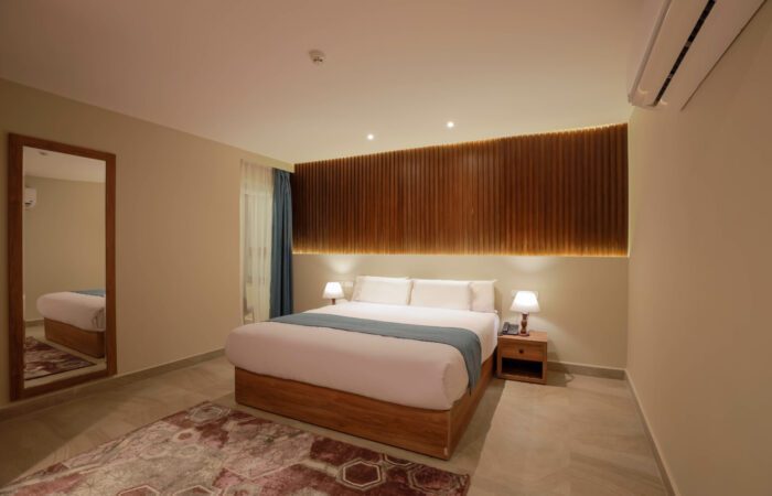 Modern hotel room with a large bed, wooden slat headboard, bedside lamps, and a full-length mirror