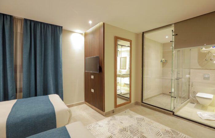 Modern hotel room with twin beds, wall-mounted TV, and a glass-enclosed bathroom