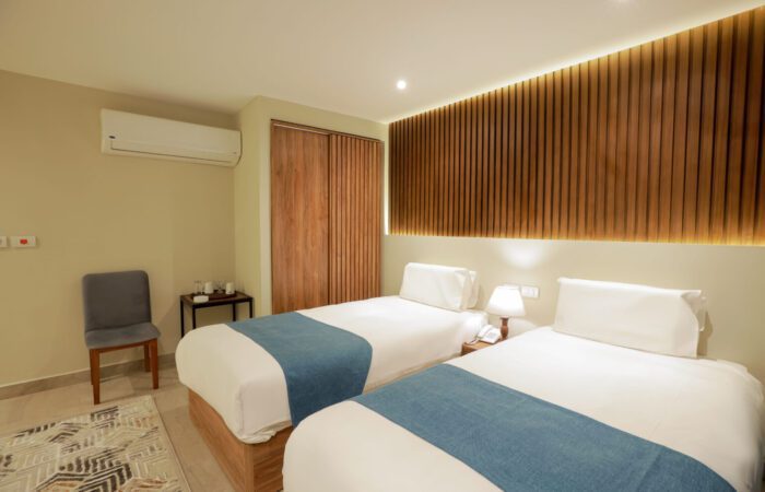 Modern hotel room with two twin beds, blue bed runners, a wooden slat wall, and air conditioning