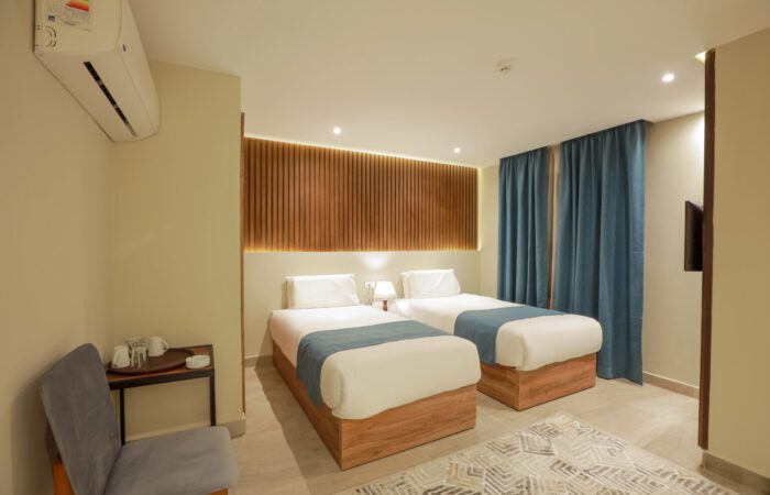 Modern hotel room featuring two twin beds with blue runners, a wooden headboard, and blue curtains