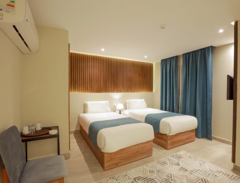 Modern hotel room featuring two twin beds with blue runners, a wooden headboard, and blue curtains