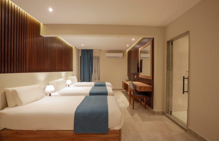 Modern hotel room with three single beds, a desk area, and a glass-enclosed bathroom