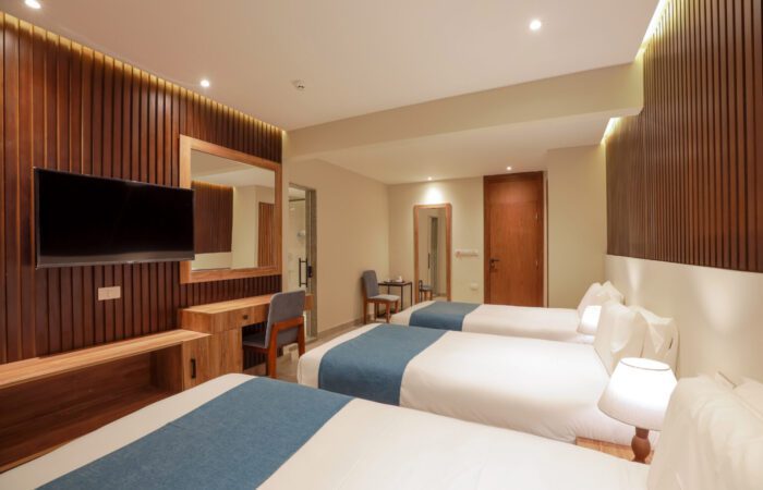 Modern hotel room featuring three single beds with blue runners, a wall-mounted TV, and a wooden desk