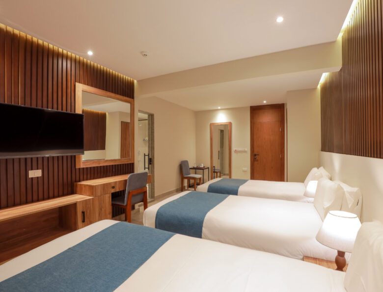 Modern hotel room featuring three single beds with blue runners, a wall-mounted TV, and a wooden desk