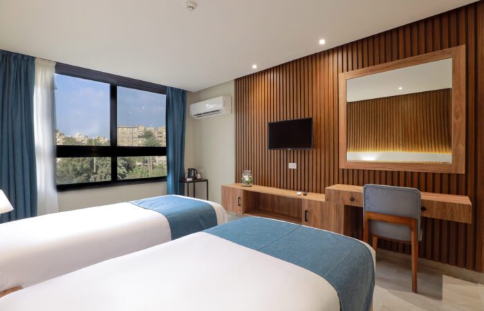 Modern hotel room with two twin beds, wooden slat wall with TV and mirror, and window with city view
