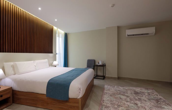 Modern hotel bedroom with a large bed, wooden slatted headboard, blue accents, and an air conditioning unit