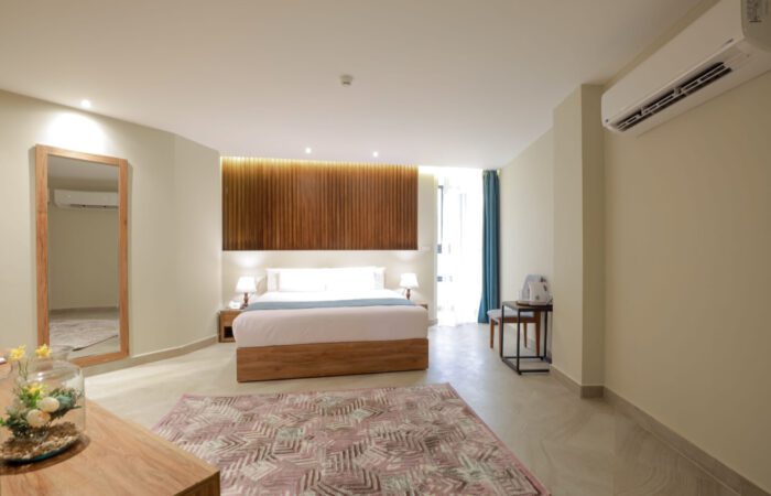 Modern hotel room with a large bed, wooden slat headboard, full-length mirror, and patterned rug