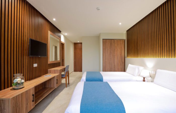 Modern hotel room featuring two twin beds with blue runners, wooden slat walls, a wall-mounted TV, and a desk