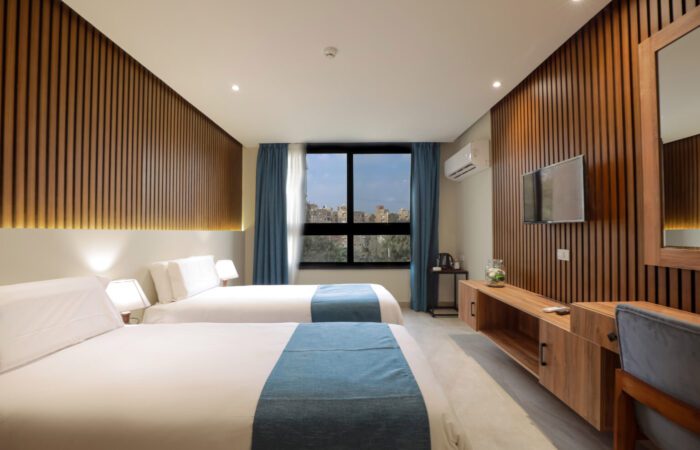 Two twin beds in a modern room with wood-paneled walls, a wall-mounted TV, and a window with a city view