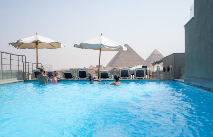 People relaxing in a rooftop swimming pool with a clear view of the Pyramids of Giza in the background
